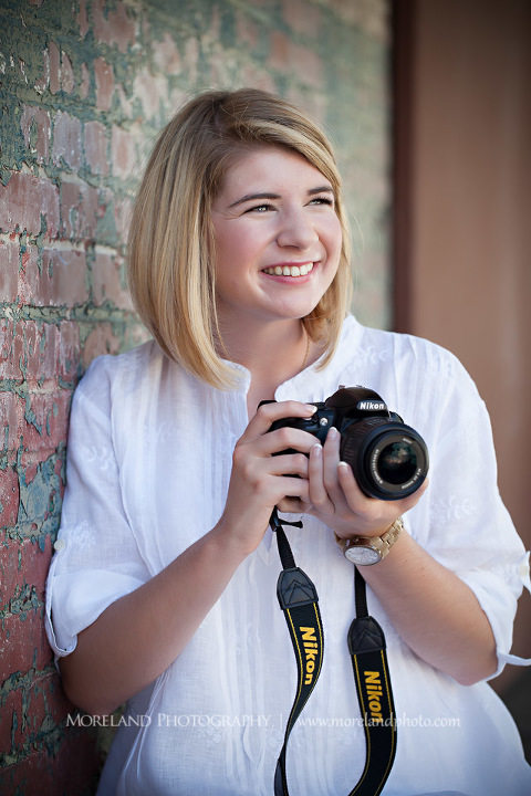 High school senior smiling as she leans against a brick wall holding her camera, Roswell Senior Photography, Atlanta Senior Portraits, Senior Photography, Atlanta Senior Photography, Moreland Photography, Mike Moreland, Class of 2016, mikemoreland, morelandphoto, excitement, class of 2016, country girl, sparkling future, big grin, posing by wall, white blouse, long shot portrait, photographer