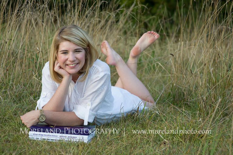 High school senior laying in a tall grass field and resting her arm on some books, Roswell Senior Photography, Atlanta Senior Portraits, Senior Photography, Atlanta Senior Photography, Moreland Photography, Mike Moreland, Class of 2016, mikemoreland, morelandphoto, excitement, class of 2016, country girl, sparkling future, big grin, classy and sassy, model portrait, white blouse, afternoon, yearbook editor, having fun, sitting in the field, bookworm, barefoot