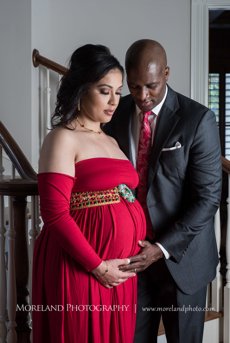 Husband in dark grey suit with red paisley tie standing next to staircase cradling wife in red maternity dress' pregnant belly, elegant maternity, pregnancy, interracial couple, romantic pregnancy photo shoot, red maternity dress, dark grey suit, red paisley tie, Atlanta area photography, Atlanta area maternity photographers, fashion maternity shoots, regal estates for photos, mike moreland photography