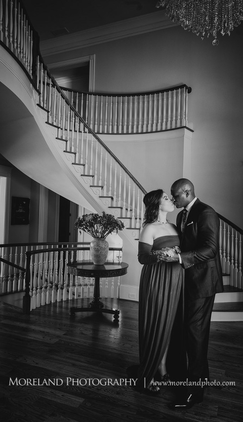 Gray scale image of sophisticated interracial couple kissing in the middle of an elegant room with chandelier next to a spiral staircase, pregnancy, interracial couple, romantic pregnancy photo shoot, red maternity dress, dark grey suit, red paisley tie, chandelier, romantic photo shoot, Atlanta area photography, Atlanta area maternity photographers, fashion maternity shoots, regal estates for photos, mike moreland photography 