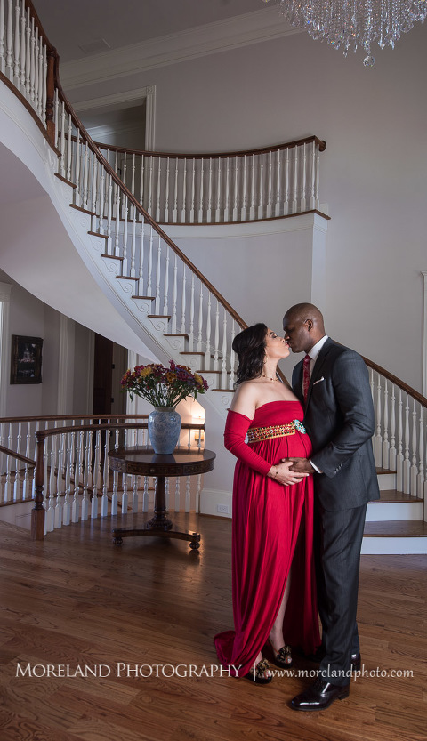Sophisticated interracial couple kissing in the middle of an elegant room with chandelier next to a spiral staircase, pregnancy, interracial couple, romantic pregnancy photo shoot, red maternity dress, dark grey suit, red paisley tie, chandelier, romantic photo shoot, Atlanta area photography, Atlanta area maternity photographers, fashion maternity shoots, regal estates for photos, mike moreland photography 