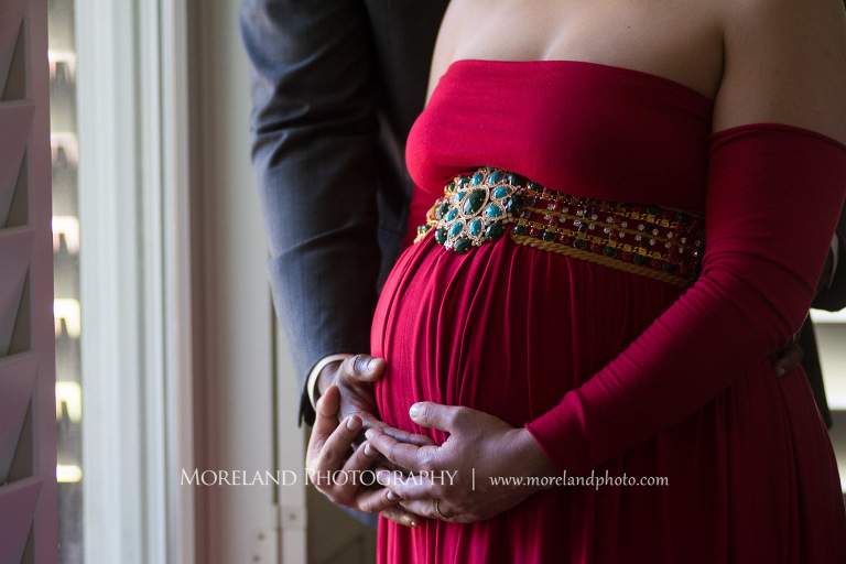 Close up image of husband in dark gray suit cradling pregnant belly of wife in red maternity dress next to a window, elegant maternity, pregnancy, interracial couple, romantic pregnancy photo shoot, red maternity dress, dark grey suit, romantic photo shoot, Atlanta area photography, Atlanta area maternity photographers, fashion maternity shoots, regal estates for photos, mike moreland photography 