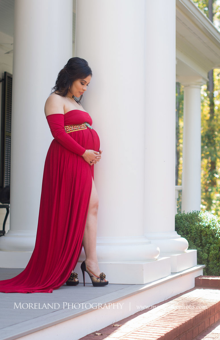 Sophisticated pregnant woman in a long red maternity dress and high heels cradling pregnant belly standing on a large porch next to big white pillars, elegant maternity, pregnancy, romantic pregnancy photo shoot, outdoor maternity photo shoot, red maternity dress, Atlanta area photography, Atlanta area maternity photographers, fashion maternity shoots, regal estates for photos, mike moreland photography 