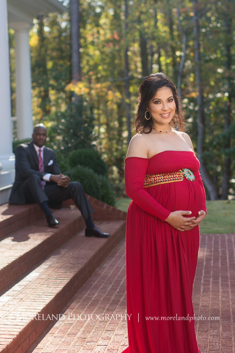 Regal pregnant woman outside in long red maternity dress smiling at camera with sophisticated husband in dark gray suit and red paisley tie sitting on steps of large porch with white pillars behind her, pregnancy, interracial couple, romantic pregnancy photo shoot, red maternity dress, dark grey suit, red paisley tie, outdoor maternity photo shoot, romantic photo shoot, Atlanta area photography, Atlanta area maternity photographers, fashion maternity shoots, regal estates for photos, mike moreland photography 