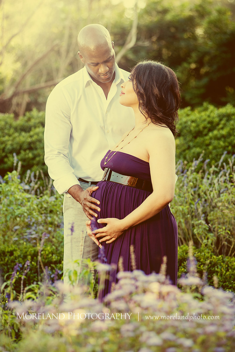 Interracial couple both holding wife in purple maternity dress' pregnant belly while standing in the middle of a garden outside, Interracial couple kissing in the middle of a garden while cradling wife's pregnant belly, pregnancy, interracial couple, outdoor pregnancy photo shoots, maternity shoots with flowers, romantic pregnancy photo shoot, purple maternity dress, outdoor maternity photo shoot, romantic moment, romantic photo shoot, Atlanta area photography, Atlanta area maternity photographers, fashion maternity shoots, regal estates for photos, mike moreland photography 