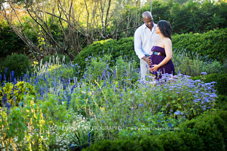 Long shot of interracial couple both holding wife in purple maternity dress' pregnant belly while standing in the middle of a garden outside, pregnancy, interracial couple, outdoor pregnancy photo shoots, maternity shoots with flowers, romantic pregnancy photo shoot, purple maternity dress, outdoor maternity photo shoot, romantic moment, romantic photo shoot, Atlanta area photography, Atlanta area maternity photographers, fashion maternity shoots, regal estates for photos, mike moreland photography 