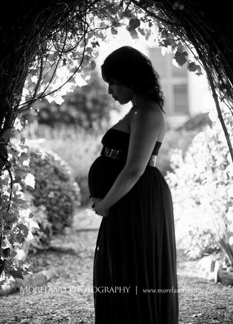 Gray scale image of pregnant woman holding belly staring downward under and arch with flowers outside, elegant maternity, pregnancy, outdoor pregnancy photo shoots, maternity shoots with flowers, romantic pregnancy photo shoot, purple maternity dress, outdoor maternity photo shoot, romantic photo shoot, Atlanta area photography, Atlanta area maternity photographers, fashion maternity shoots, regal estates for photos, mike moreland photography 