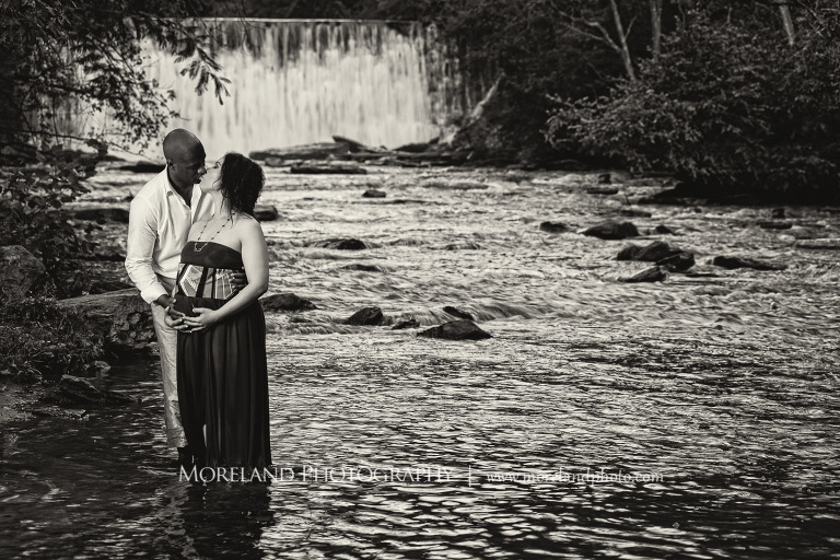 Gray scale image of interracial couple kissing in the middle of a river with a waterfall behind them, pregnancy, interracial couple, outdoor pregnancy photo shoots, maternity shoots with nature, fun maternity shoots, romantic pregnancy photo shoot, purple maternity dress, outdoor maternity photo shoot, romantic moment, romantic photo shoot, maternity photo shoots with water, Atlanta area photography, Atlanta area maternity photographers, fashion maternity shoots, mike moreland photography 