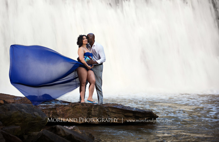 husband kissing pregnant wife in a flowing purple maternity dress' cheek while caressing her belly on a rock in front of a gorgeous waterfall, pregnancy, fun maternity shoots, outdoor pregnancy photo shoots, maternity shoots with nature, waterfall, purple maternity dress, romantic photo shoot, interracial couple, Atlanta area photography, Atlanta area maternity photographers, fashion maternity shoots, regal estates for photos, mike moreland photography 