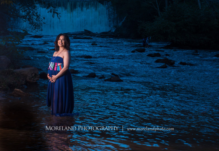 Dark lit image of a beautiful pregnant woman in a purple maternity dress smiling at the camera in the middle of a river with a waterfall behind her, husband kissing pregnant wife in a flowing purple maternity dress' cheek while caressing her belly on a rock in front of a gorgeous waterfall, pregnancy, fun maternity shoots, outdoor pregnancy photo shoots, maternity shoots with nature, waterfall, purple maternity dress, romantic photo shoot, Atlanta area photography, Atlanta area maternity photographers, fashion maternity shoots, regal estates for photos, mike moreland photography 