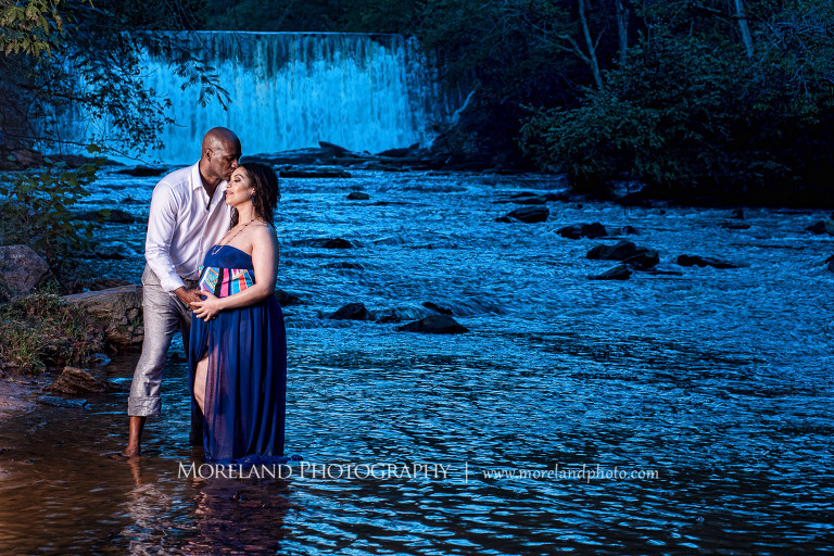 Dark lit image of a husband kissing pregnant wife in a flowing purple maternity dress' forehead while caressing her belly in the middle of a river in front of a gorgeous waterfall, pregnancy, fun maternity shoots, outdoor pregnancy photo shoots, maternity shoots with nature, waterfall, purple maternity dress, romantic photo shoot, interracial couple, Atlanta area photography, Atlanta area maternity photographers, fashion maternity shoots, regal estates for photos, mike moreland photography 