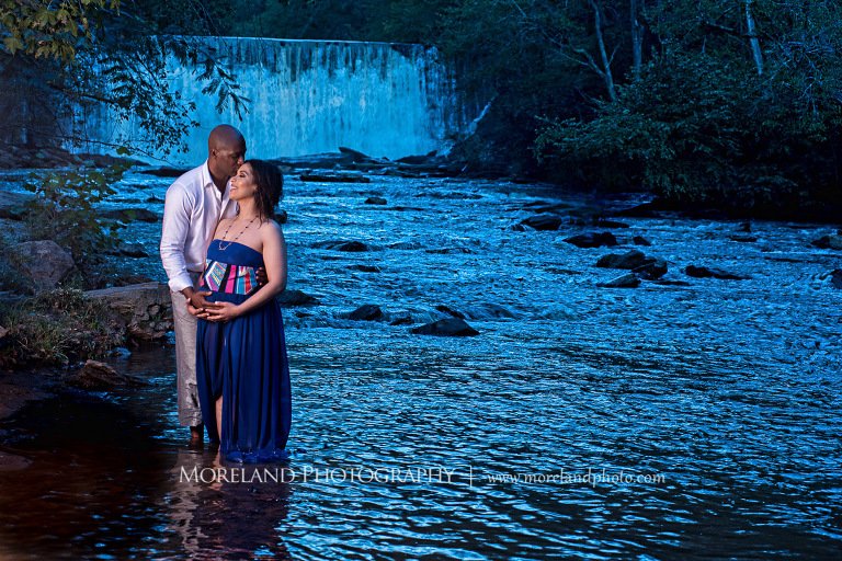 Dark lit image of a husband kissing smiling pregnant wife in a flowing purple maternity dress' forehead while caressing her belly in the middle of a river in front of a gorgeous waterfall, pregnancy, fun maternity shoots, outdoor pregnancy photo shoots, maternity shoots with nature, waterfall, purple maternity dress, romantic photo shoot, interracial couple, Atlanta area photography, Atlanta area maternity photographers, fashion maternity shoots, regal estates for photos, mike moreland photography 