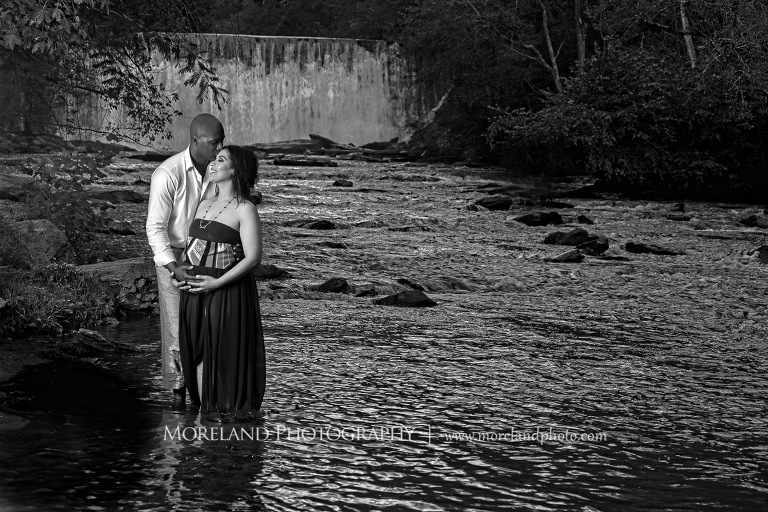 Gray-scale image of a husband kissing smiling pregnant wife in a flowing purple maternity dress' forehead while caressing her belly in the middle of a river in front of a gorgeous waterfall, pregnancy, fun maternity shoots, outdoor pregnancy photo shoots, maternity shoots with nature, waterfall, purple maternity dress, romantic photo shoot, interracial couple, Atlanta area photography, Atlanta area maternity photographers, fashion maternity shoots, regal estates for photos, mike moreland photography 