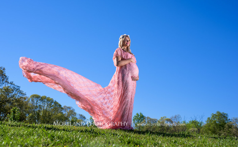 Pregnant woman standing in a field in a pink dress as she holds her stomach and her dress flows in the wind, Atlanta Maternity Photographer, Maternity Photos Atlanta, Georgia Maternity Photography, Moreland Photography, Mike Moreland, Outdoor Maternity, Maternity on the River, 