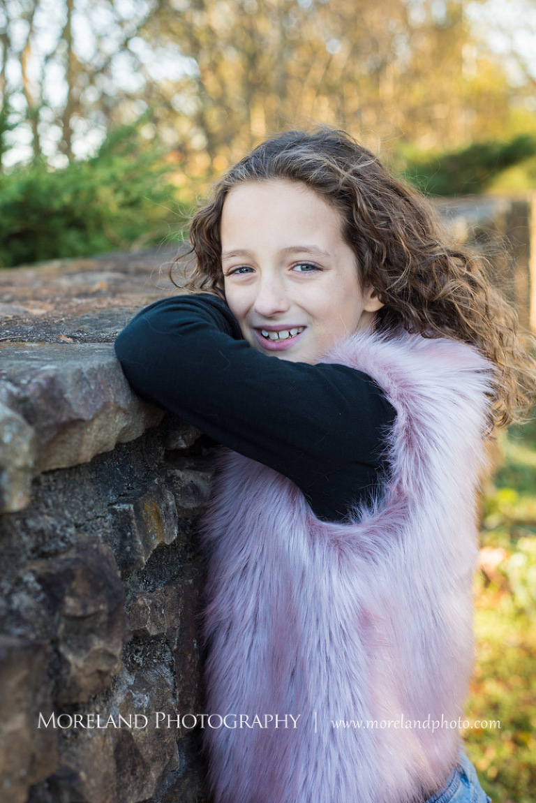 Portrait of little girl looking over her arm at camera leaning against brick wall outside during daytime in pink furry vest and black shirt, Atlanta Portrait Photographer, Atlanta Child Photographer, Atlanta Family Photographer, Moreland Photography, Mike Moreland