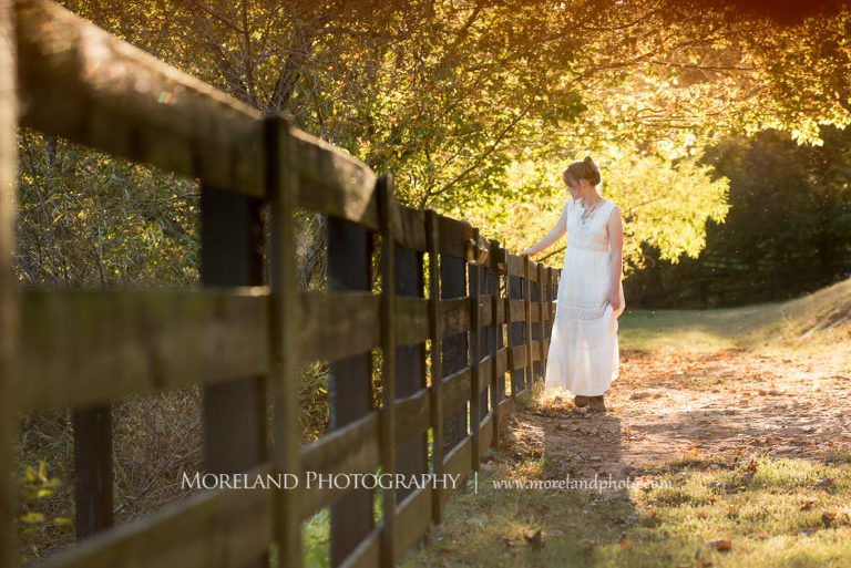 Georgia Senior portrait of a girl in nature standing by a long black wooden fence , Mike Moreland, Moreland Photography, Atlanta Portrait Photographer, Senior Photography Atlanta, Kings Ridge Christian Academy, Nature, Equestrian, Georgia Senior Portrait, Outdoors Photography, Georgia Girl, Nature, Girl Standing Wood Fence, White Dress, Wooden Back Fence, Fall, Leaves Changing, Stable 
