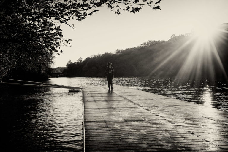 black and white portrait of boy walking down dock with sun flare from the right, creative black and white portrait, documentary photography, teenager by water